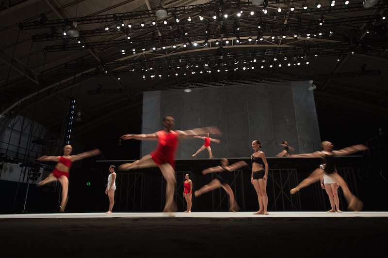 Several dancers in red jump in arabesque while others stand still